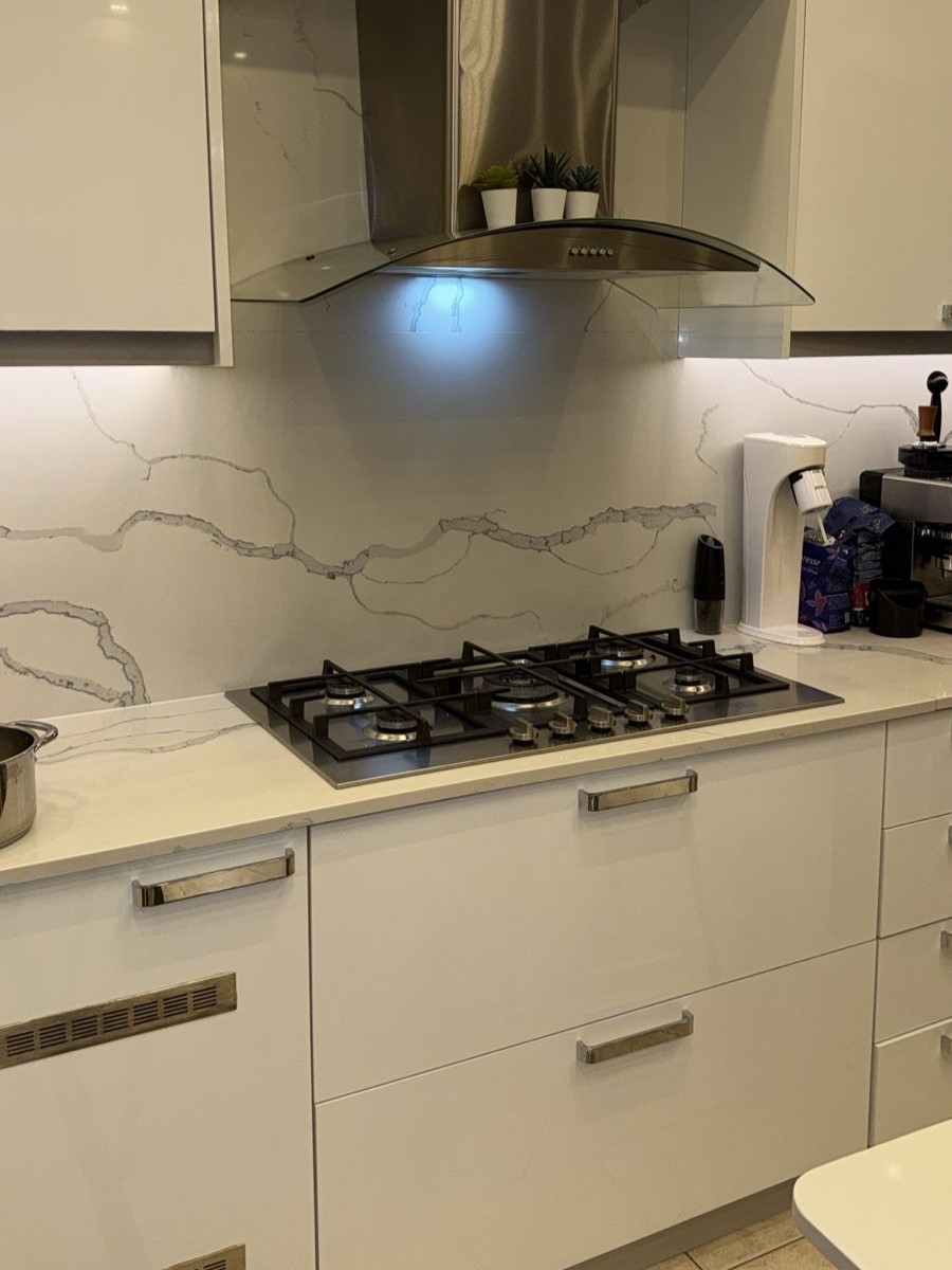 0a5bf298-4682-444b-8a91-9df207a5840d White modern kitchen with a five-burner gas stove, marble-veined backsplash, and a curved stainless range hood; small potted plants sit on the hood.
