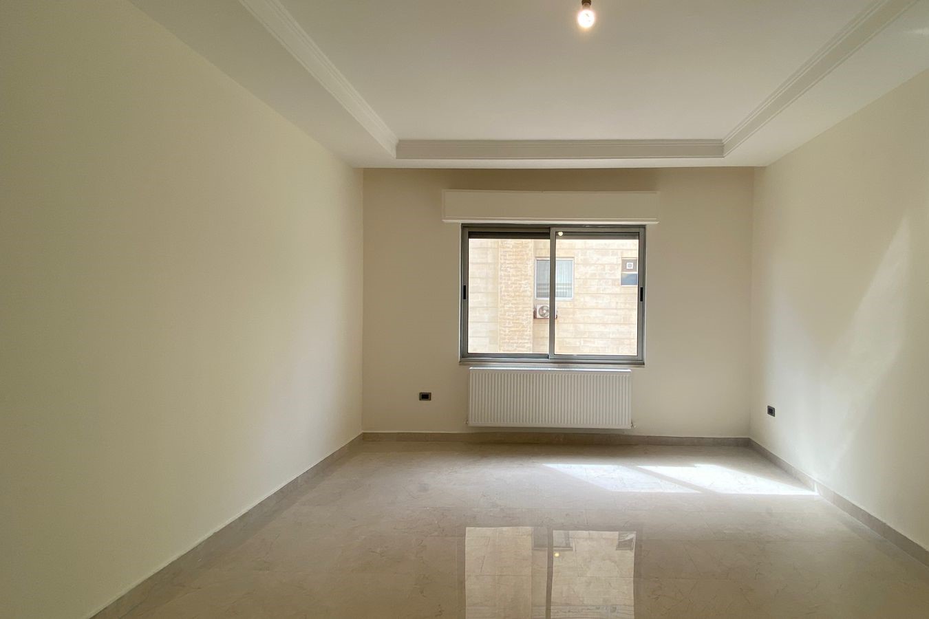 0a847f5d-b829-4a02-8463-721de9f7b7b0 Empty rectangular room with pale beige walls, a window centered on the far wall above a white radiator, and a glossy tiled floor illuminated by daylight from outside.