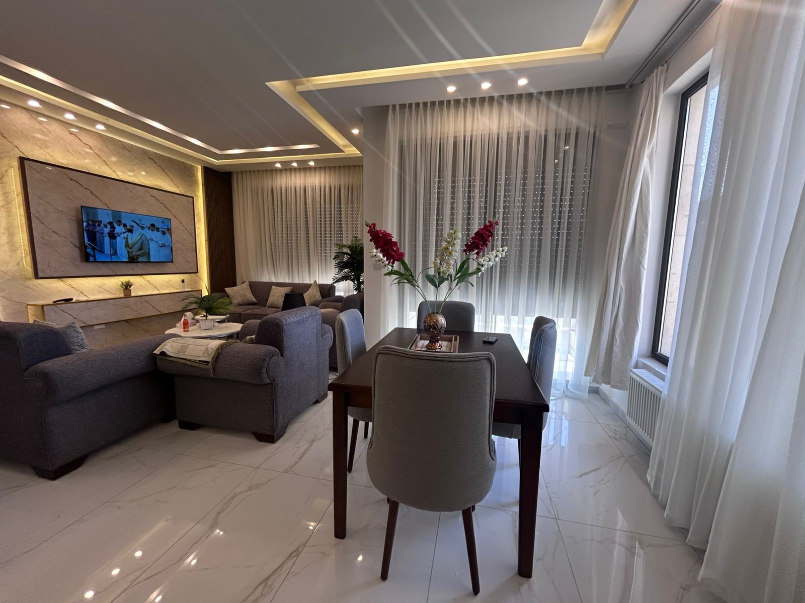 0ab326ea-fbcd-4f2f-8744-e102bf096f74 Modern living room with gray sofas, marble floor, and a dining table with a vase of red and white flowers.