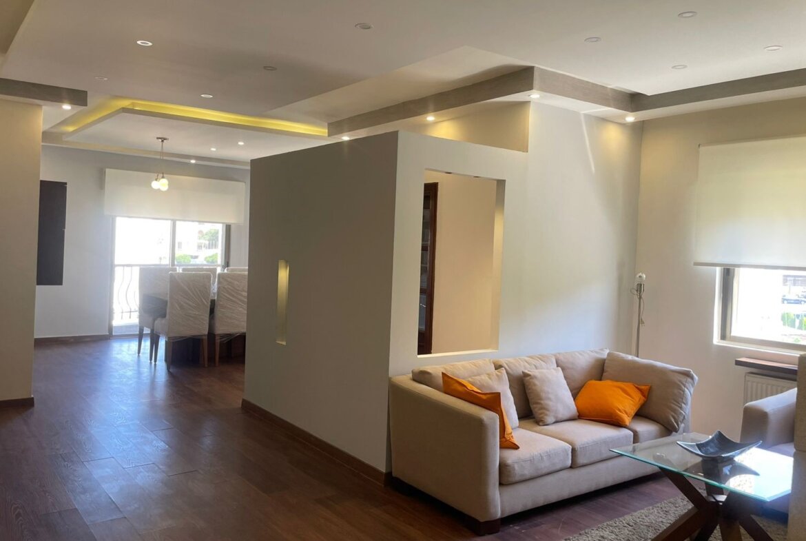 Open-concept living area with beige sofa and orange pillows, glass coffee table, and wooden floors; dining area in background near large window.