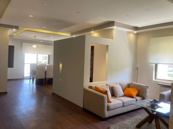 Open-concept living area with beige sofa and orange pillows, glass coffee table, and wooden floors; dining area in background near large window.