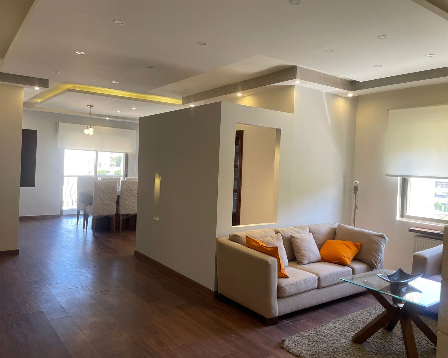 0ae8c8e3-8f98-4ebc-9bd5-d53c75d5073c Open-concept living area with beige sofa and orange pillows, glass coffee table, and wooden floors; dining area in background near large window.