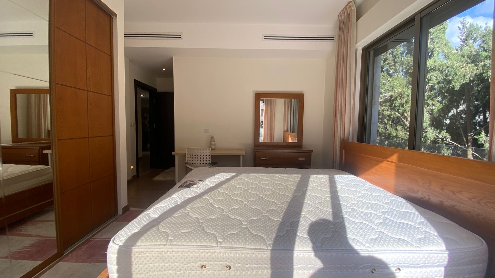 0b7e138b-e0a7-4b04-aa40-6f2ac378481b Bright bedroom with a large window showing green trees, a wooden bed frame and a white mattress, and a dresser with mirror opposite the window.