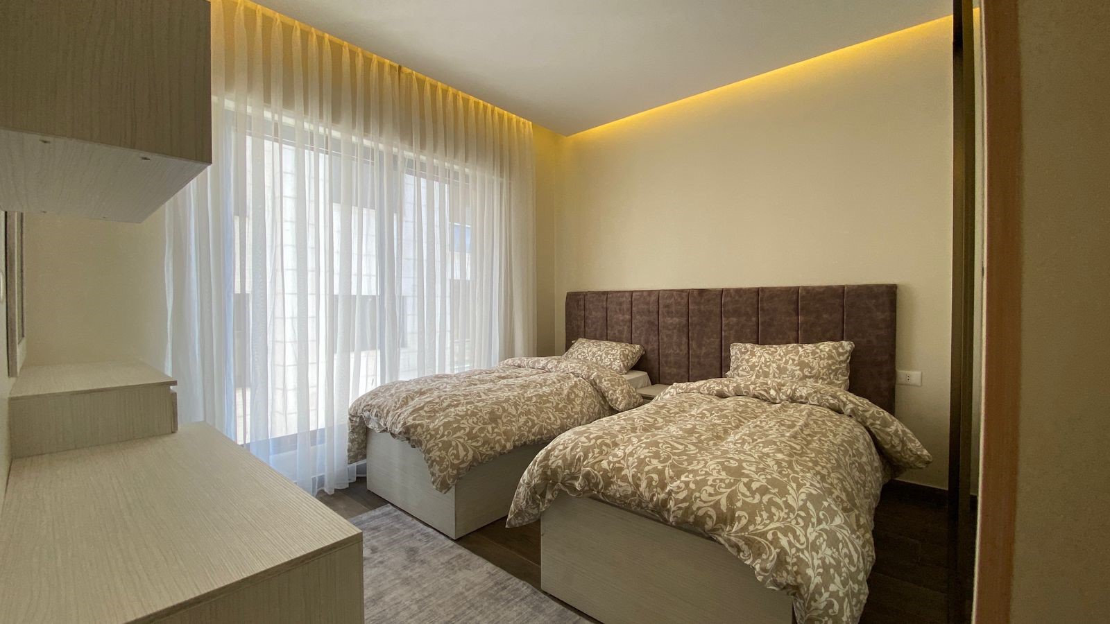 0bb67799-bc88-4f32-9b69-69cdb05e0902 Twin bedroom with a padded brown headboard, two matching patterned duvets, and pillows, next to a large window with sheer curtains.