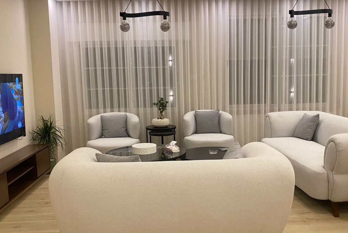 Cozy modern living room with a beige curved sofa, two armchairs, and a glass coffee table facing a TV.