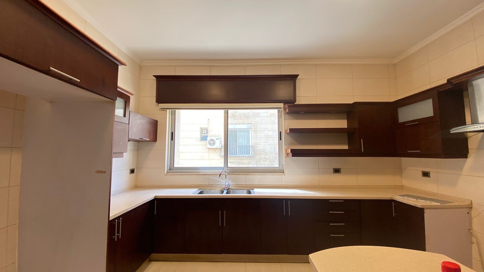 0e1f6dbd-dbe5-40df-9a4d-b30c113b3539 L-shaped kitchen with dark wood cabinets, white countertops, and a double sink under a window.