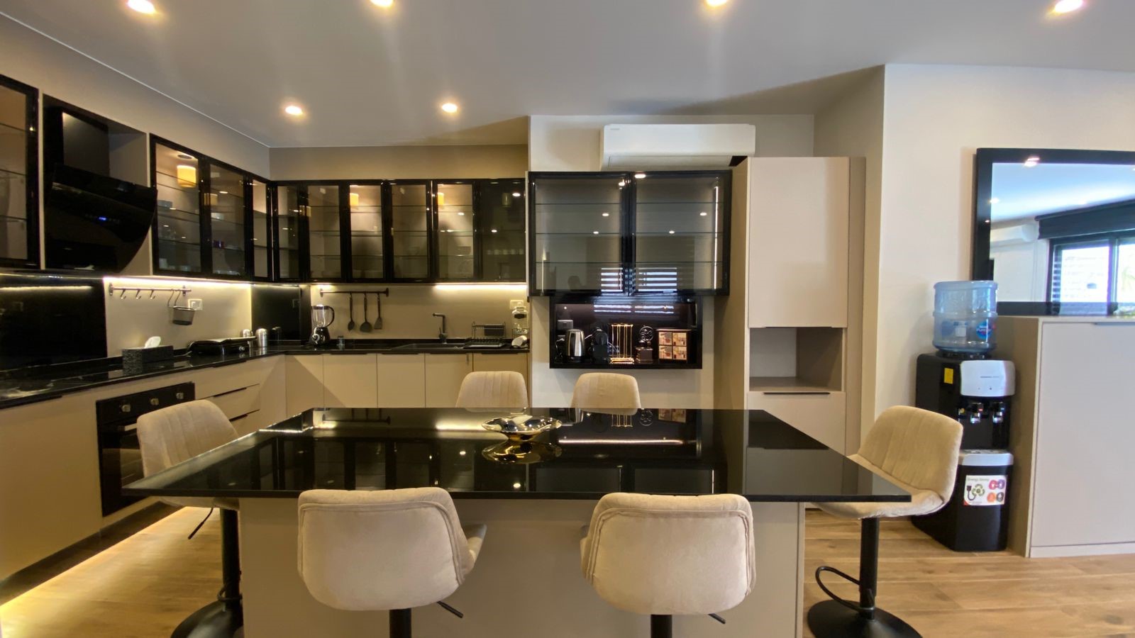 0e3671d9-a8ce-45a3-9877-3049f6089f23 Modern kitchen with black-glass cabinets, a glossy black island, beige stools, and a water cooler in the corner.