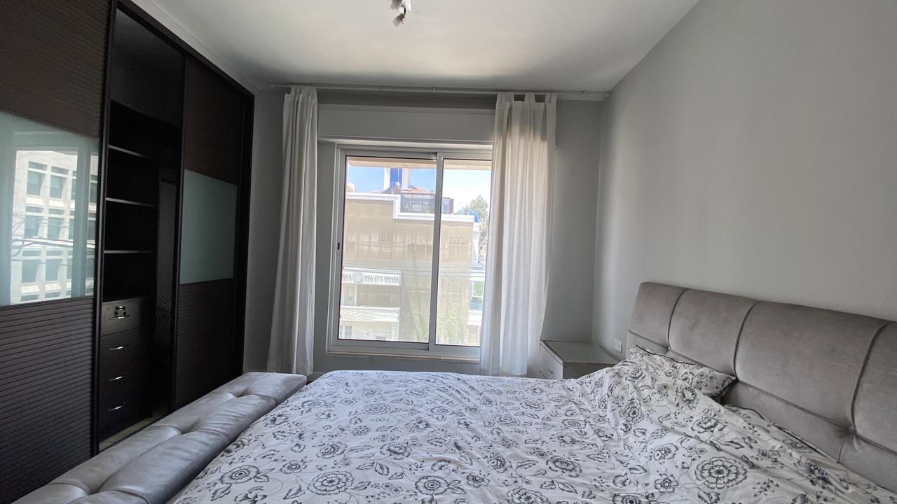 10018168-3ae5-429f-9364-c32564d07c74 A modern bedroom with a large upholstered bed, patterned bedding, and a dark wooden wardrobe along the left wall. A window with white curtains lets in light across the room.