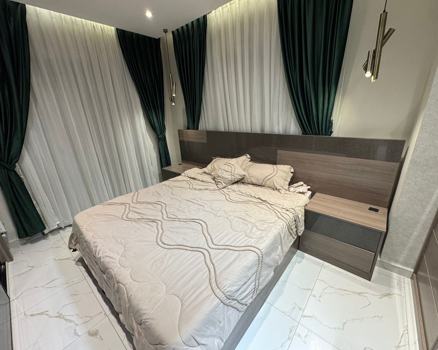 1090e711-2dc3-4dc3-ad72-24f5f28948d0 Modern bedroom with a double bed, beige quilted bedding, and matching wood headboard with two nightstands; dark green drapes and sheer white curtains.