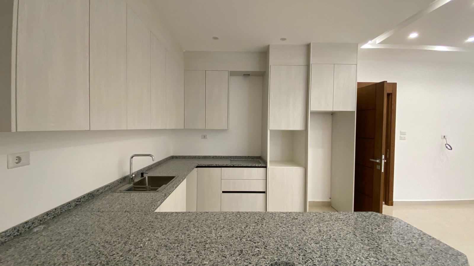 11c9c9fa-e8e2-4653-8180-a3b6d2fa33b7 Modern L-shaped kitchen with light wood cabinets, gray granite countertops, and a stainless steel sink.