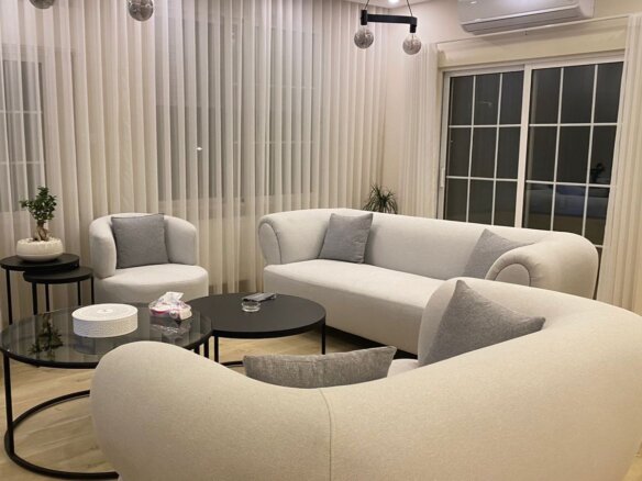 Cream curved sofa set with gray cushions around glass coffee tables in a modern living room with sheer white curtains and plants to the side, creating a neutral, cozy vibe.