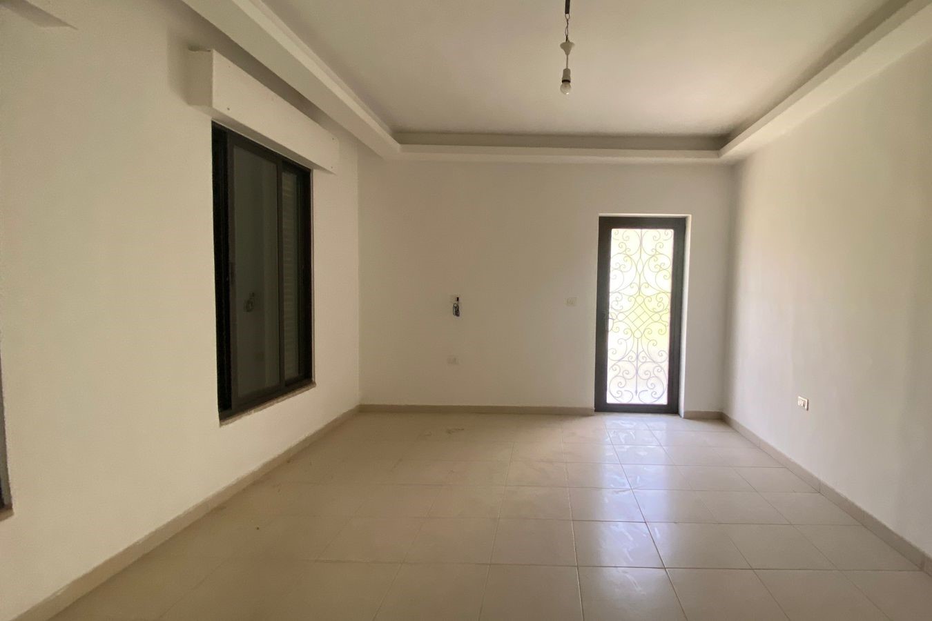 12b55045-67f8-462d-943b-476c15ccdb43 Empty, unfurnished room with white walls, tiled floor, a large window on the left, and a decorative metal door to the right, bare light bulb overhead.