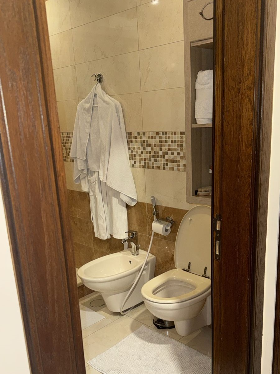 12f5aa18-cdc8-45e4-80b5-059b618b394c 1 Bathroom with white toilet and bidet, robe on hook, shelf with towels on tiled wall in background