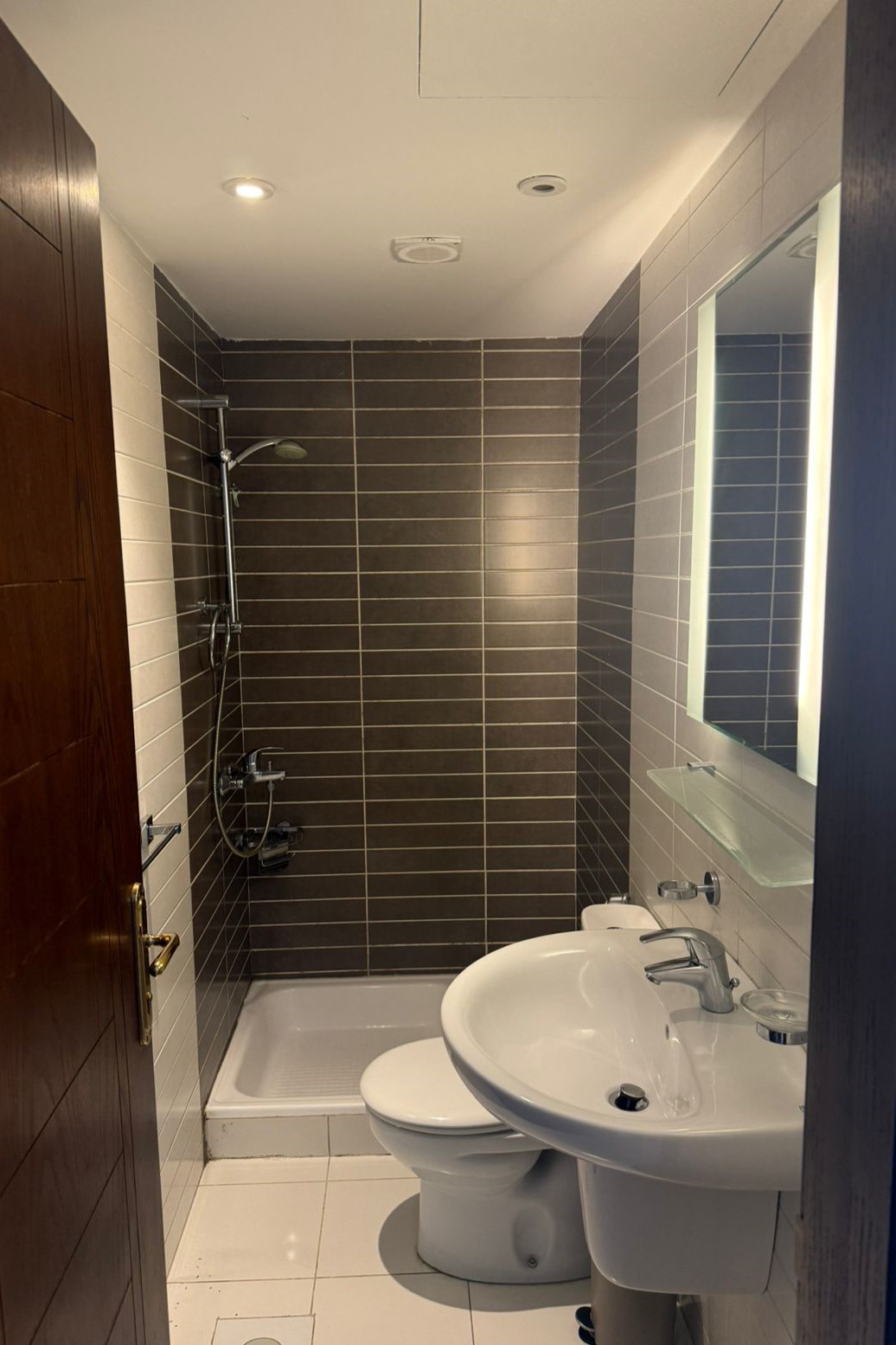13f9908c-fe14-43bb-9ea2-2eab2b4f0247 Compact bathroom with dark tile shower area at back, and white sink and toilet in foreground; wood door on left and mirror with light on right.