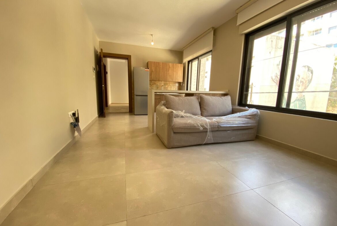 Open-plan living area with a plastic-wrapped gray sofa, light tile flooring, and large windows filling the space with daylight.
