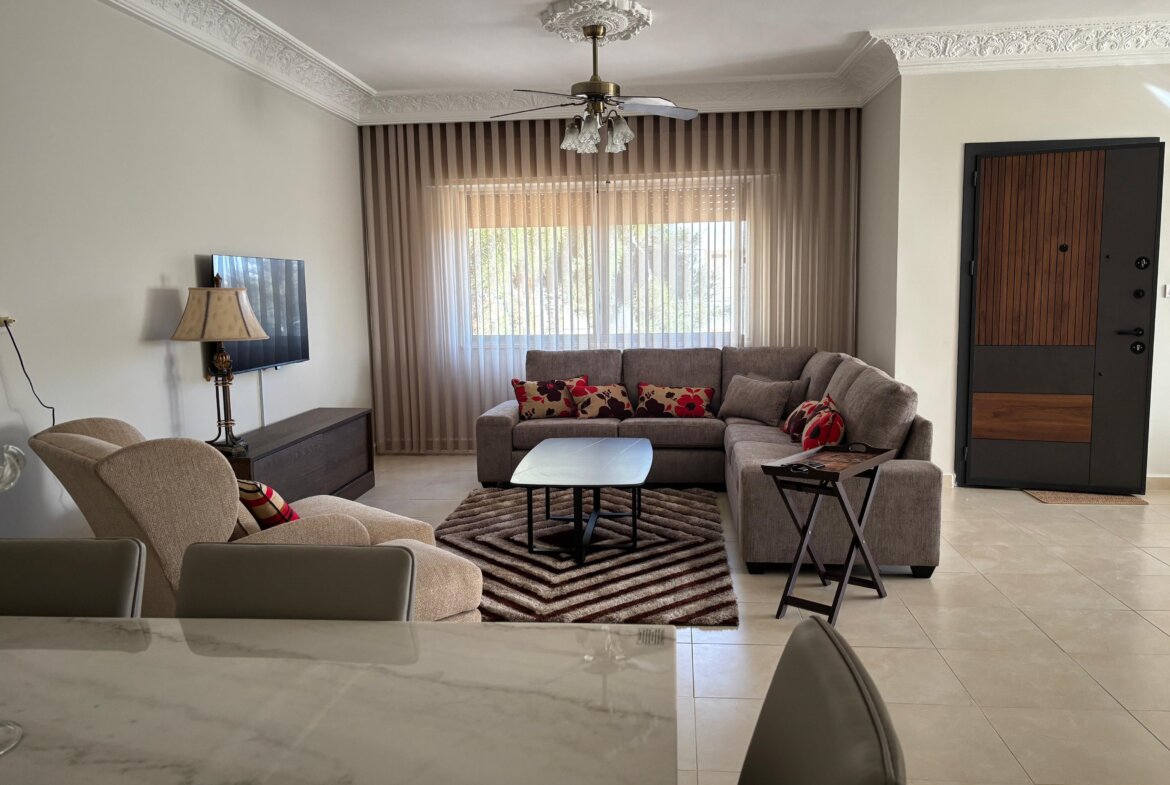 Cozy living room with an L-shaped beige sectional, red patterned cushions, and a coffee table on a striped rug near a window with vertical blinds.