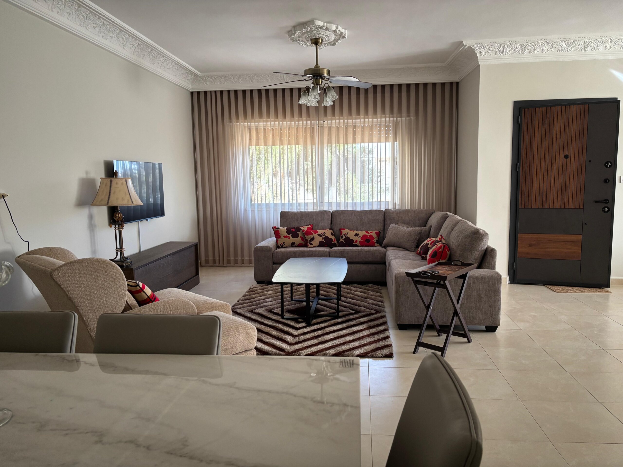 158ff4b3-72cd-4622-9ec0-f69064dd54d3 Cozy living room with an L-shaped beige sectional, red patterned cushions, and a coffee table on a striped rug near a window with vertical blinds.