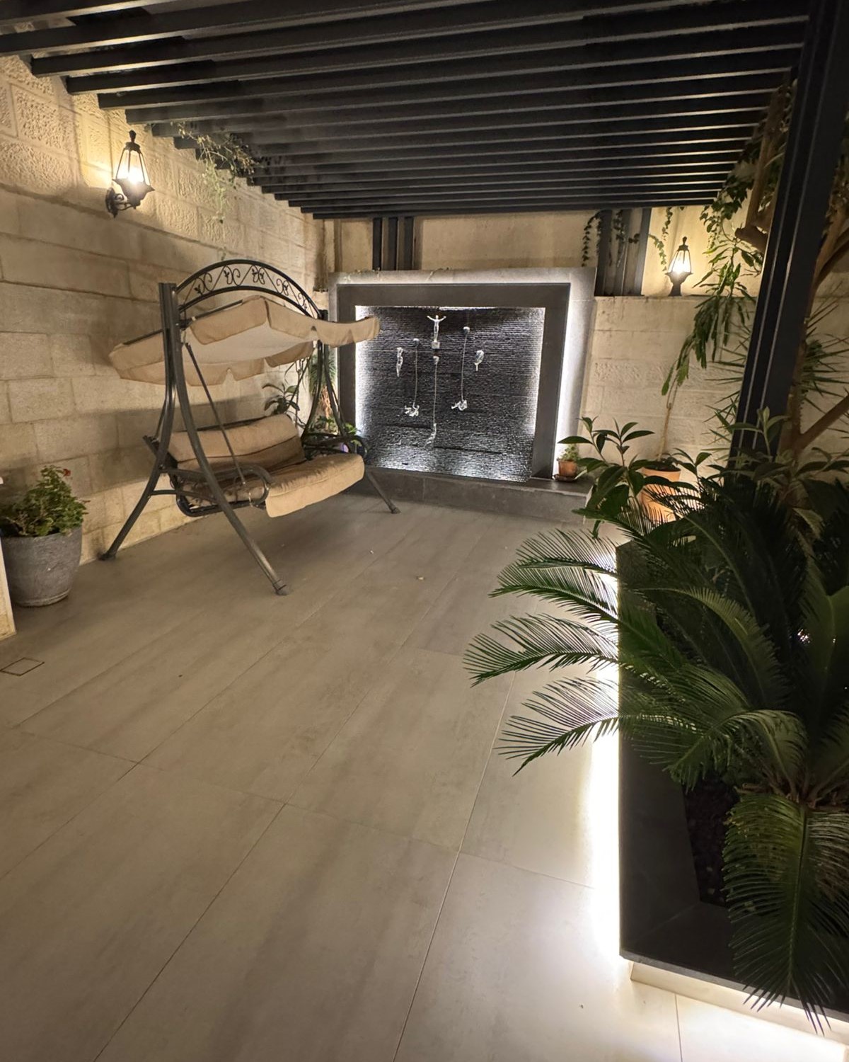 15d332a0-064f-44cb-b7e3-6196ede47b88 Cozy outdoor patio with a beige cushioned metal swing chair beside a stone wall and a water feature wall fountain in the back corner, warm lantern lights glowing.