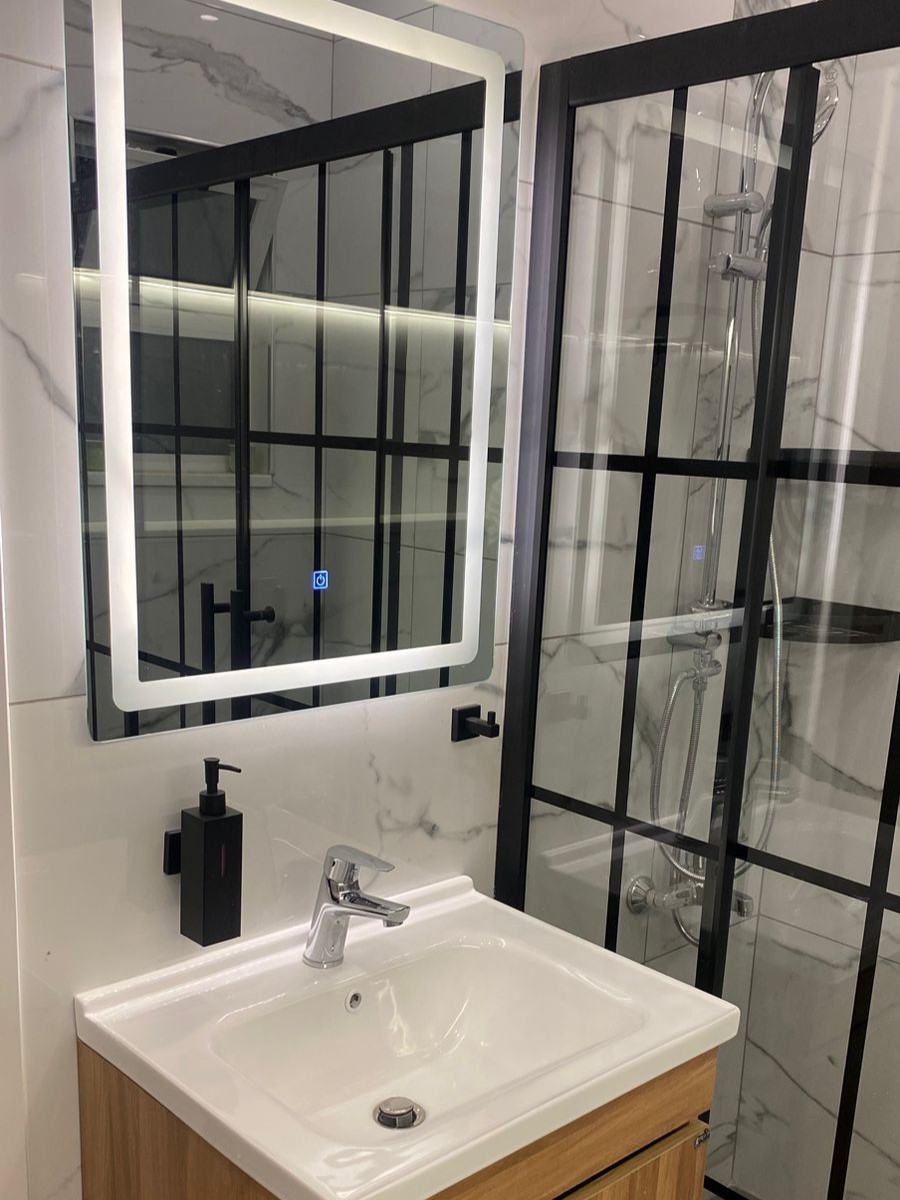 17475b8f-9f2a-480c-9bbb-574a5bd49619 Modern bathroom with LED-framed mirror above a white sink and wooden vanity, plus a black soap dispenser and a glass shower with a black frame.