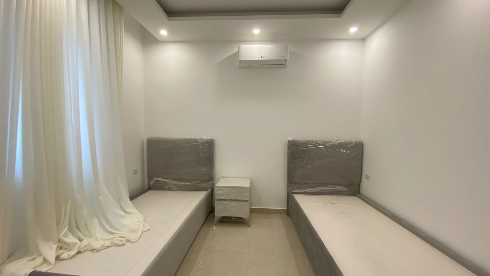 17987d5d-cadf-40ea-9cdc-08037f736b4b Minimal white bedroom with two single beds covered in plastic, a small white nightstand between them, and an air conditioner on the wall.
