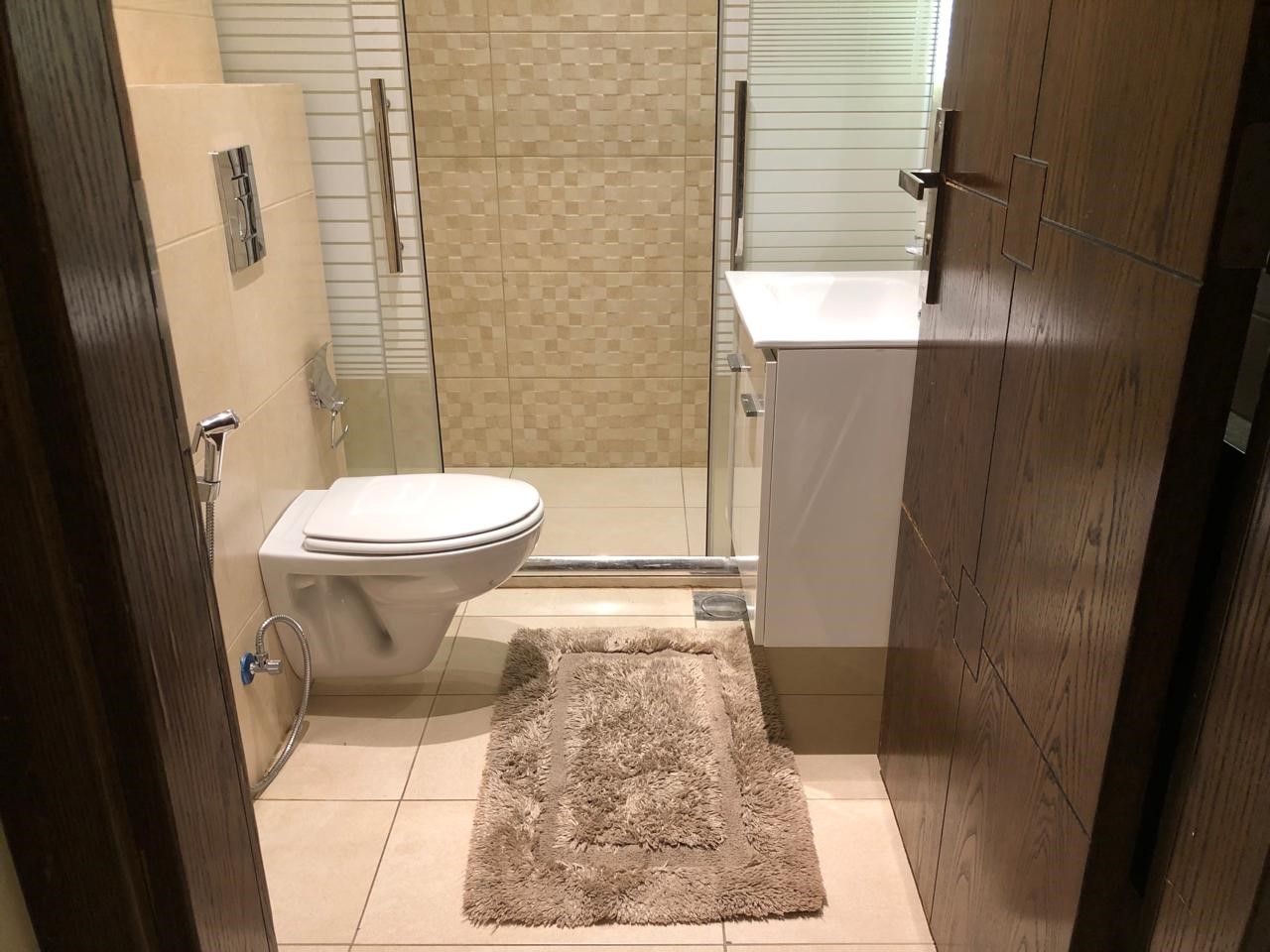 1849f1cc-e26e-4edb-879f-8dec01203ed7 Compact, modern bathroom with a wall-mounted toilet, glass shower enclosure, white vanity, and a beige shag rug on beige tile floors.
