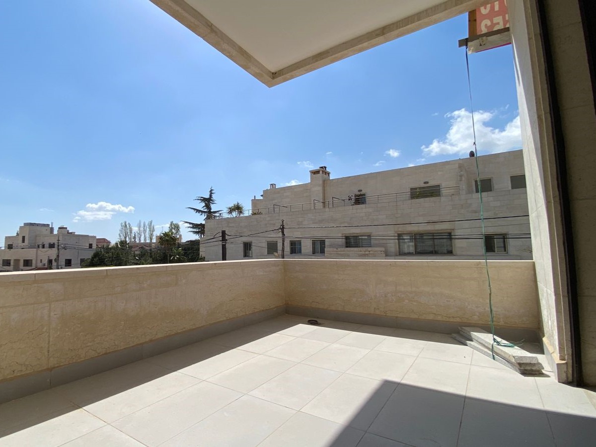 19565197-dcc8-415d-9a2e-ef63f8315cff Sunny balcony with a beige stone railing and tiled floor, overlooking white buildings under a clear blue sky.