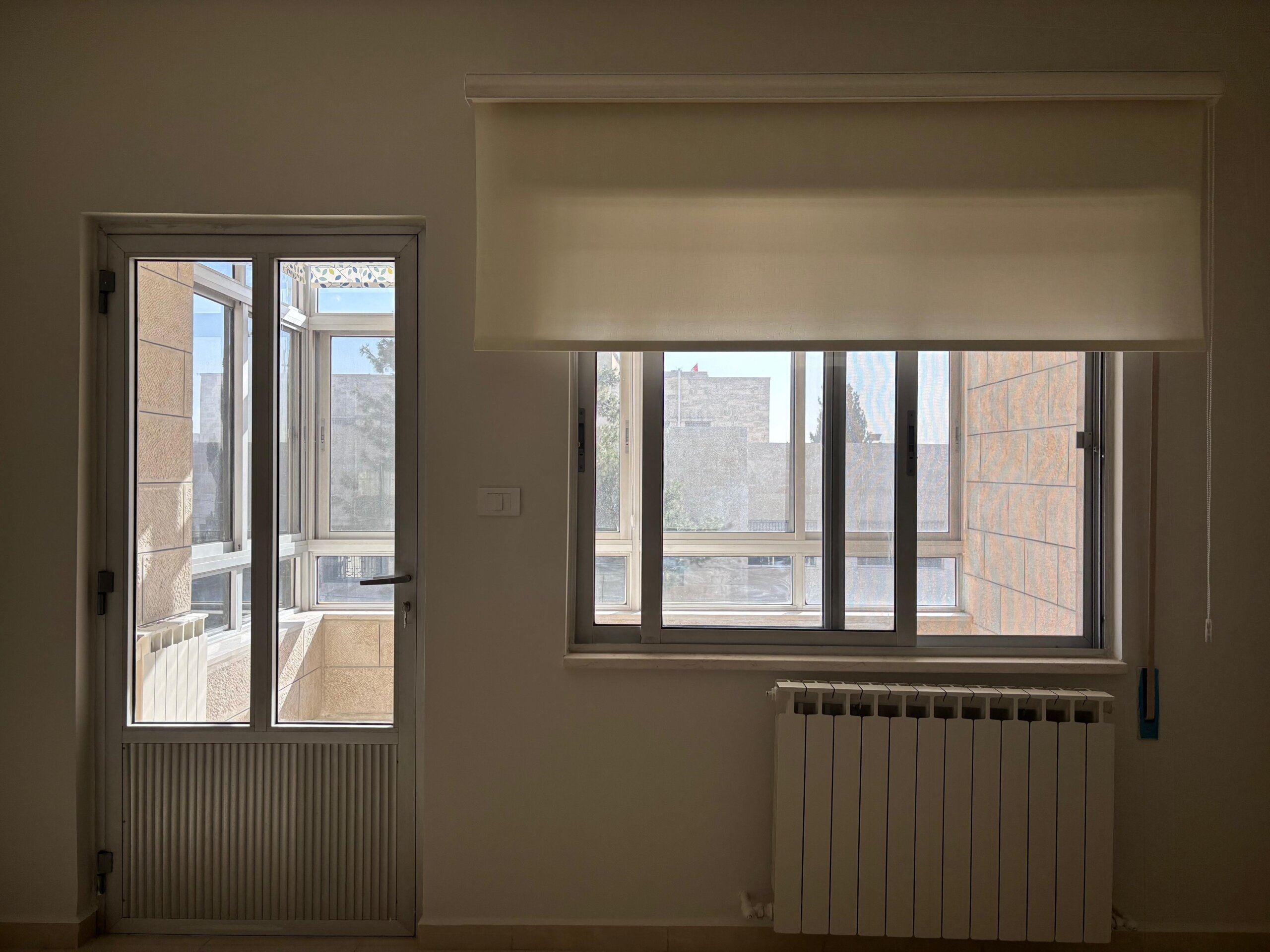 1a9fe273-af3f-44da-8df9-c03e727a72e9 Sunlit interior with a large window and a white roller blind, a radiator beneath the window, and a glass door to a balcony on the left side of the room