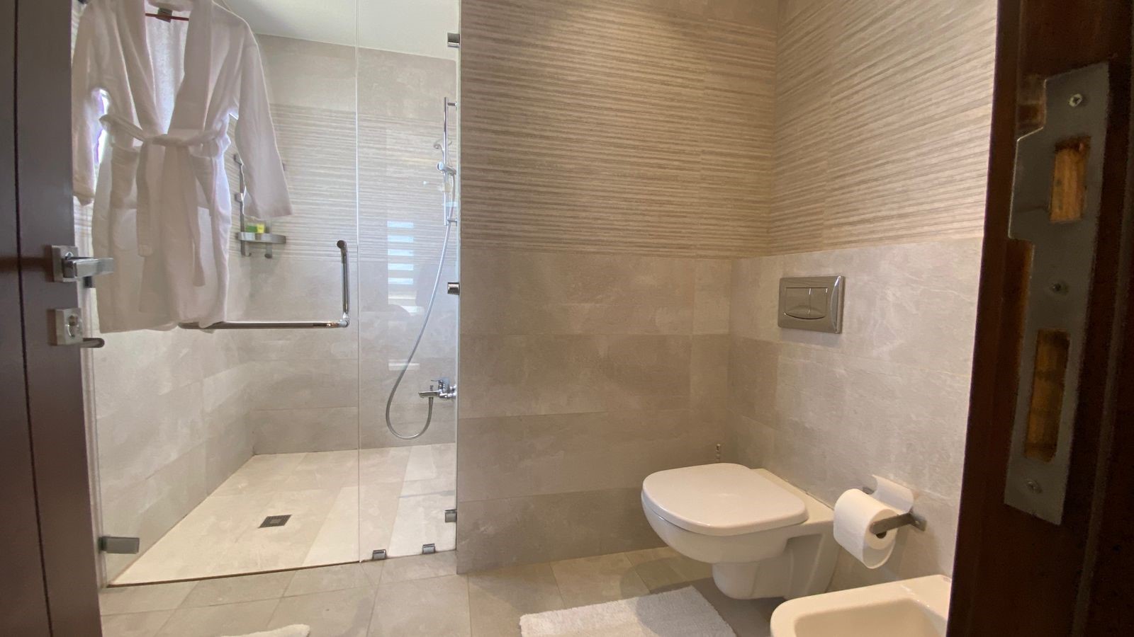 1b1afdce-366f-413e-97b3-3cce9aea77a4 Modern bathroom with glass shower enclosure, wall-mounted toilet, and a white robe hanging on the door.