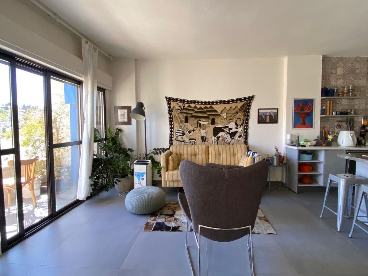 Sunlit living room with sliding glass doors to a balcony, potted plants, striped beige couch, a large wall tapestry, and a small open kitchen on the right.