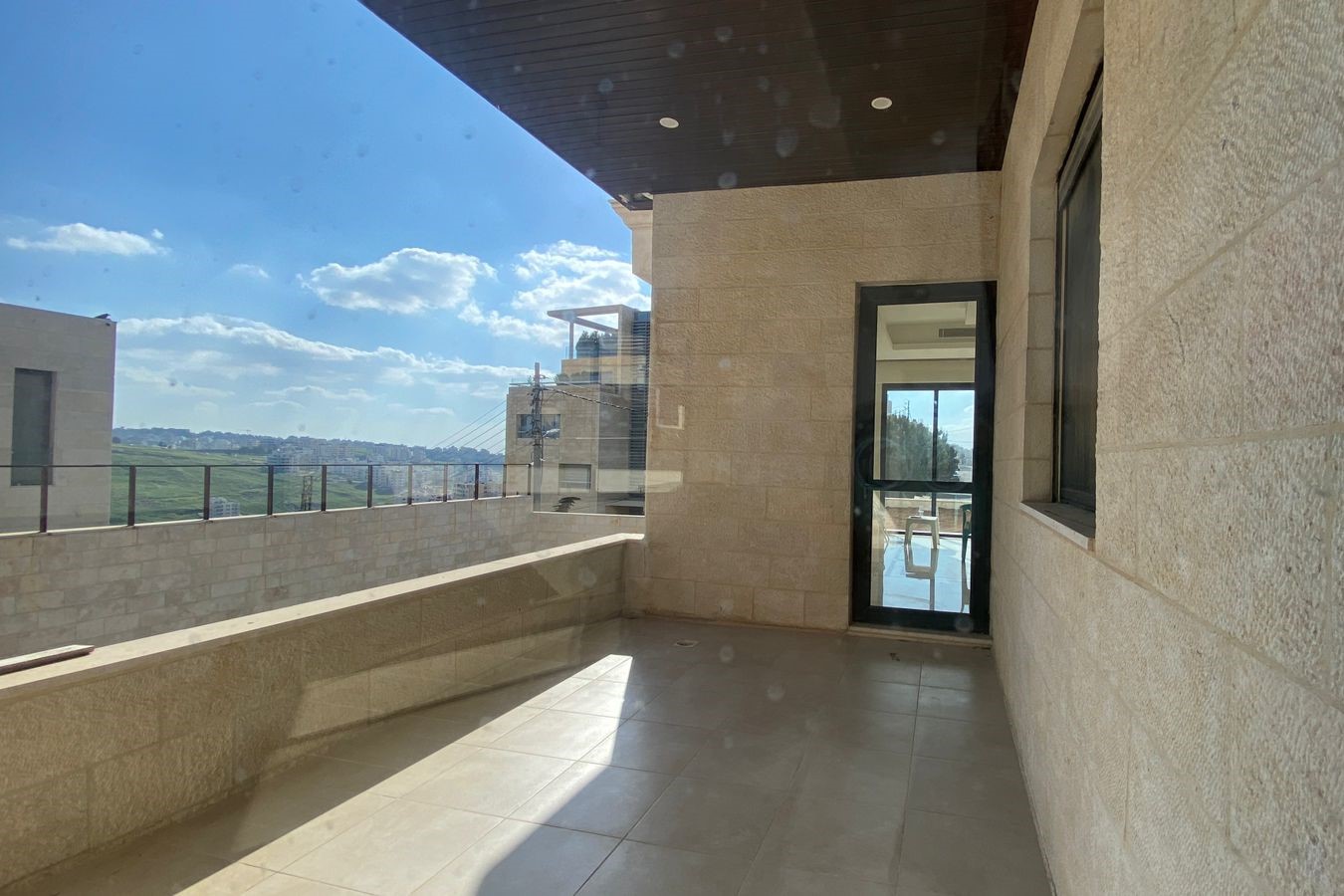1c4f9ff5-7539-465f-a969-02f14348e0c8 Sunlit balcony with beige stone walls, glass railing, and a door opening to an interior space; blue sky and distant hills in view.