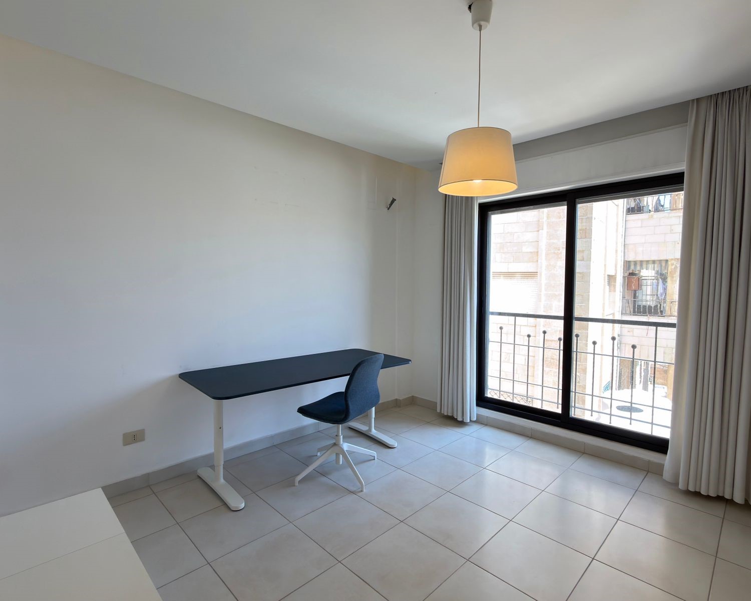 1cdeb1ed-41c0-4dc8-9e4a-f550b09ad9af A small study corner with a dark desk and blue swivel chair beside a large window with white curtains in a bright, empty room.