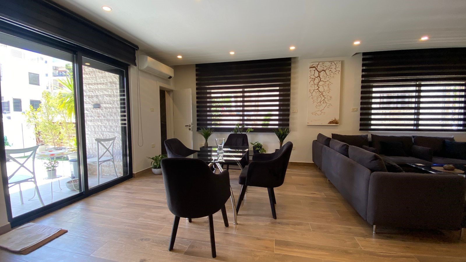 1d749bdc-b99f-4837-873d-8a5632f268f3 Open living-dining room with glass doors to a sunny patio, a black glass dining table and four black chairs, and a grey sectional sofa.
