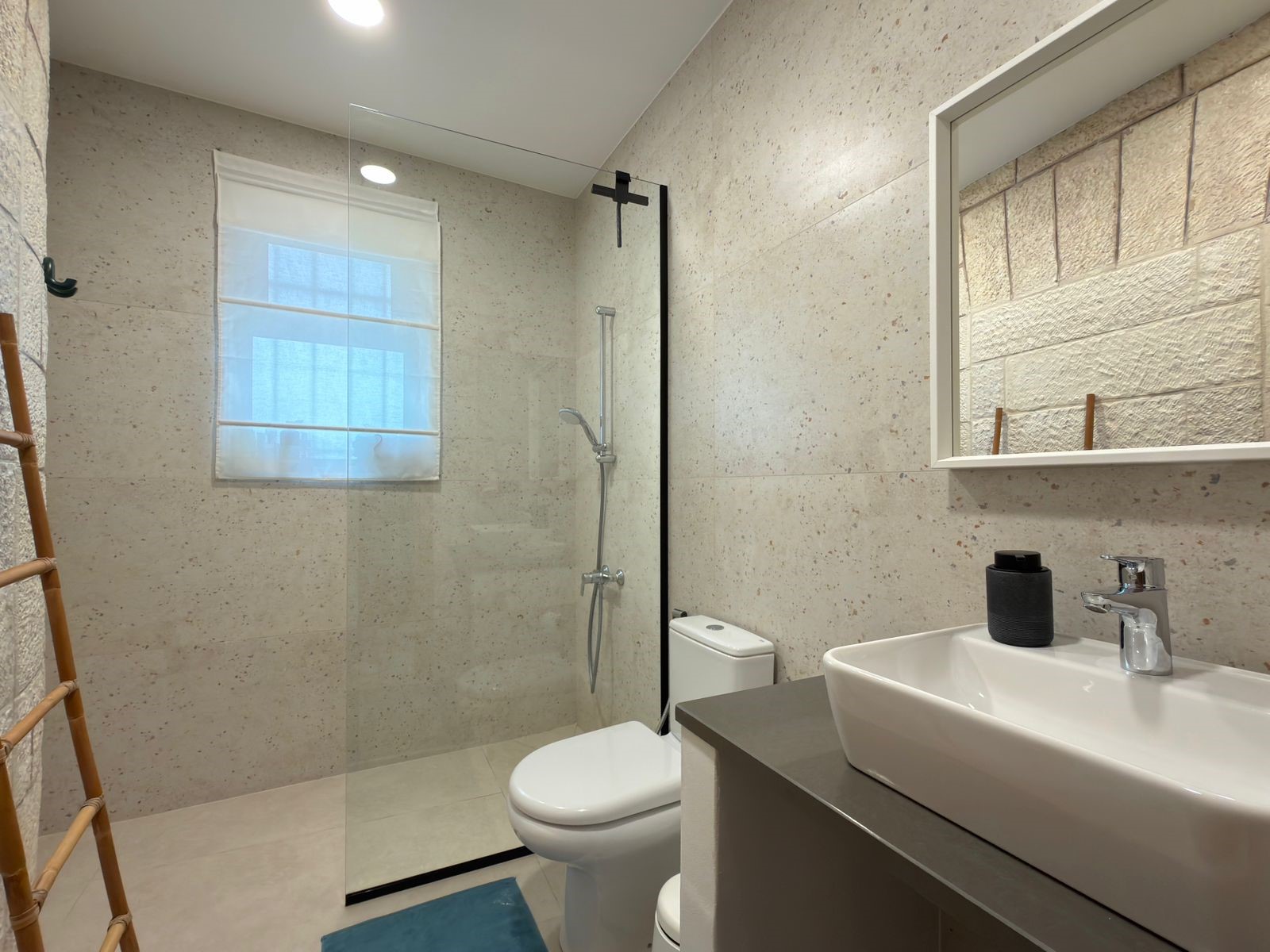 1e385cae-47e6-491c-9490-18763eb3c716 Modern bathroom with glass-walled shower, toilet, and rectangular sink; beige speckled walls and a window blind.