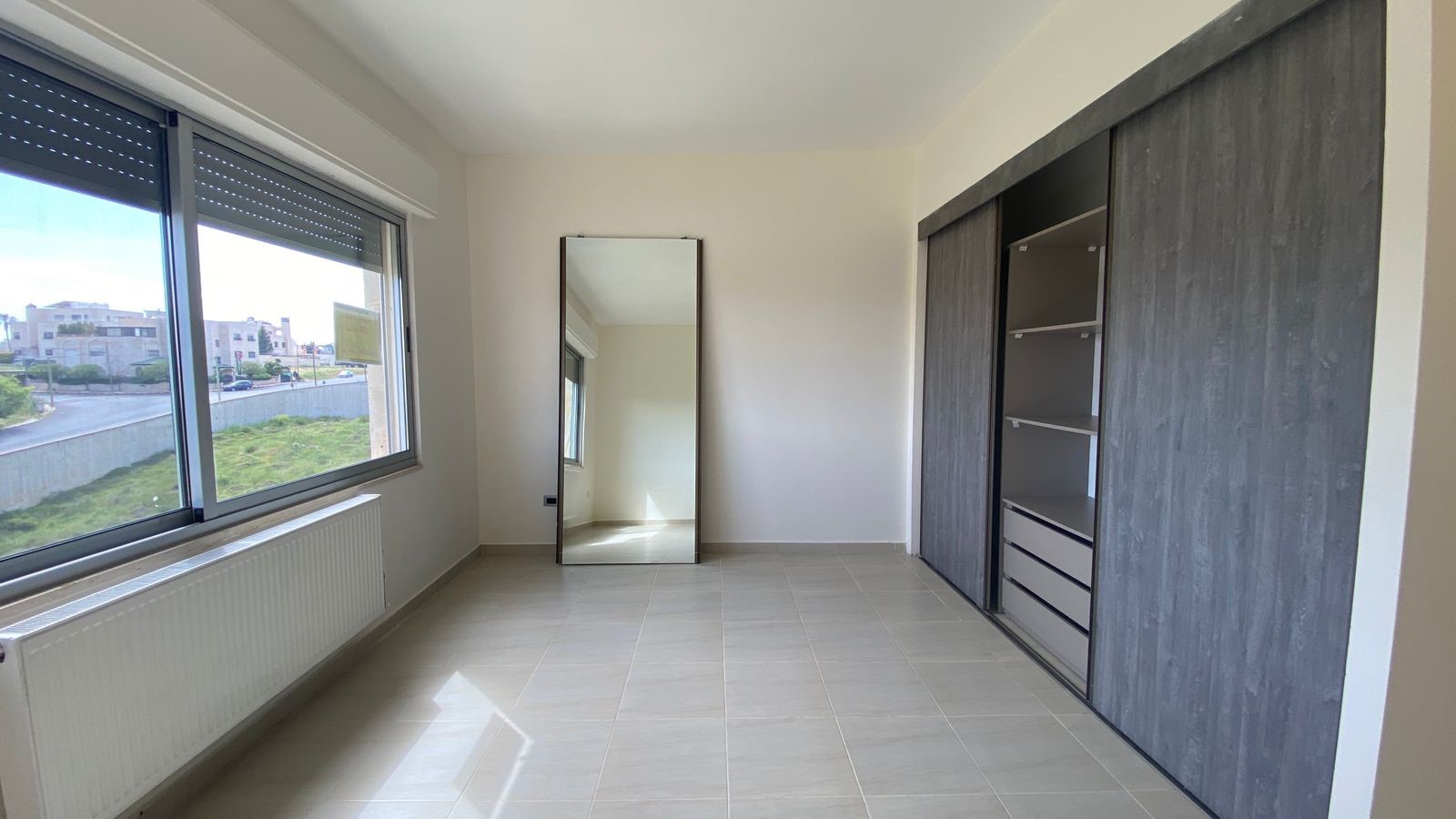 1e4cb615-0e62-4510-a2c9-7e57331490a4 Empty bedroom with large window, radiator along the wall, a full-length freestanding mirror, and a built-in dark wood wardrobe withopen shelves and drawers on the right.