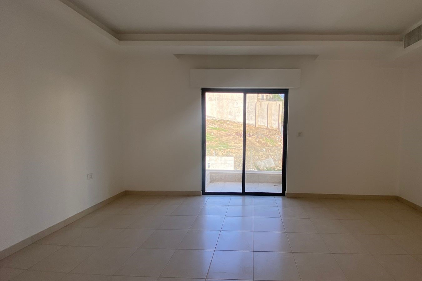 1ebc2fd8-d962-4323-b519-3768b8a03225 Empty room with tiled beige floor and a large sliding glass door opening to an outdoor patio with a hillside beyond.