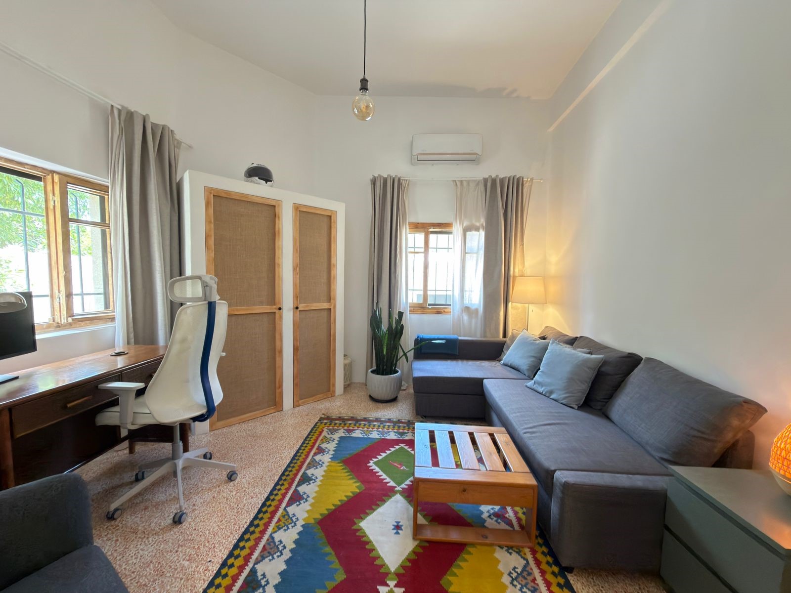 1f675b58-7025-4b1a-9723-9f030f16367b Bright living room with gray sectional sofa, colorful geometric rug, and wooden coffee table. A desk and white chair sit by a window to the left, with a tall wardrobe and potted plant nearby.