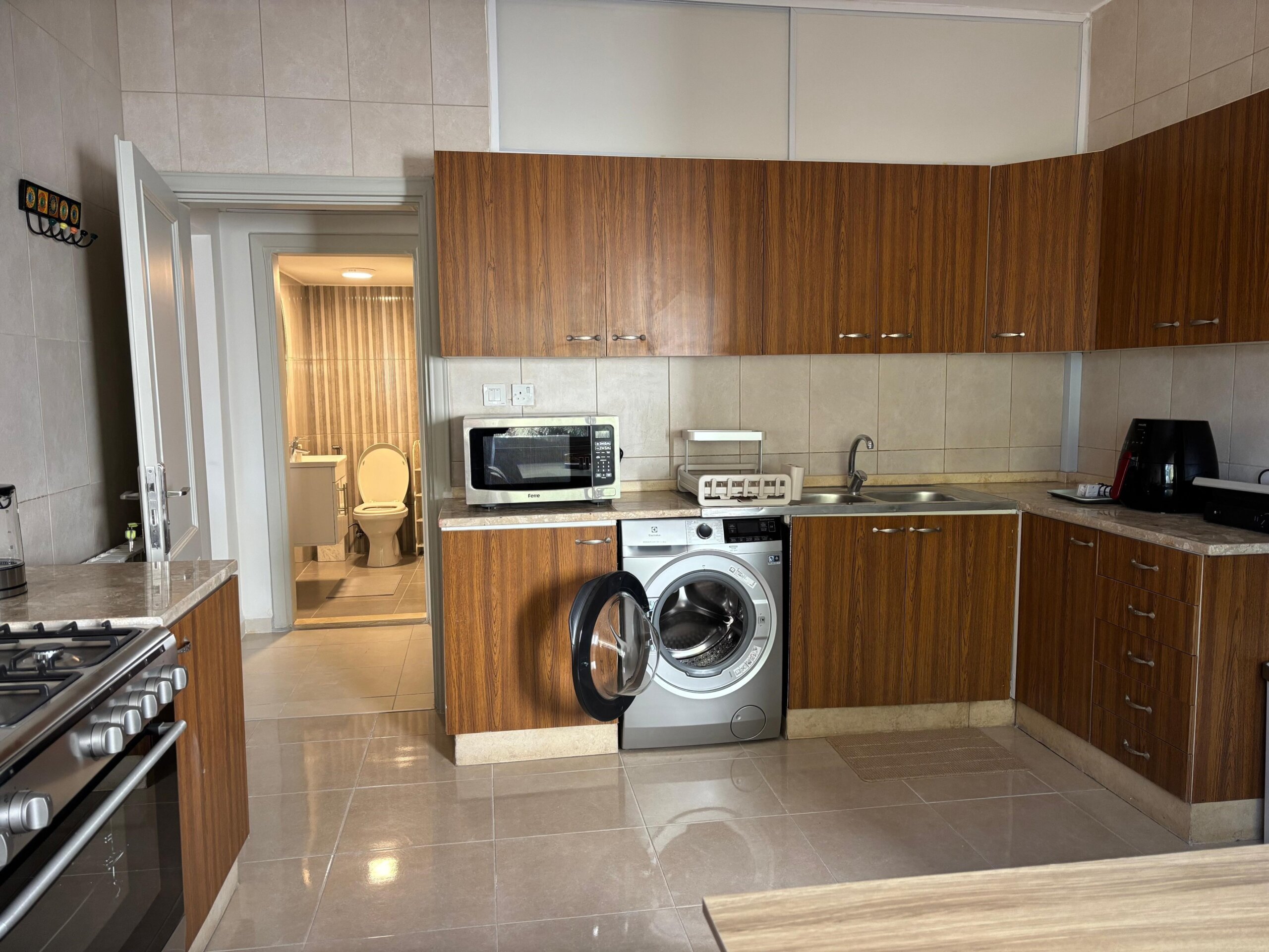 1f7d8a58-c217-4c13-82bd-7fbf10ae87fa Kitchen with wooden cabinets and beige tiled walls; foreground stove, oven, counter, and a front-loading washing machine under the counter near the sink. A doorway leads to a bathroom with a toilet in view.