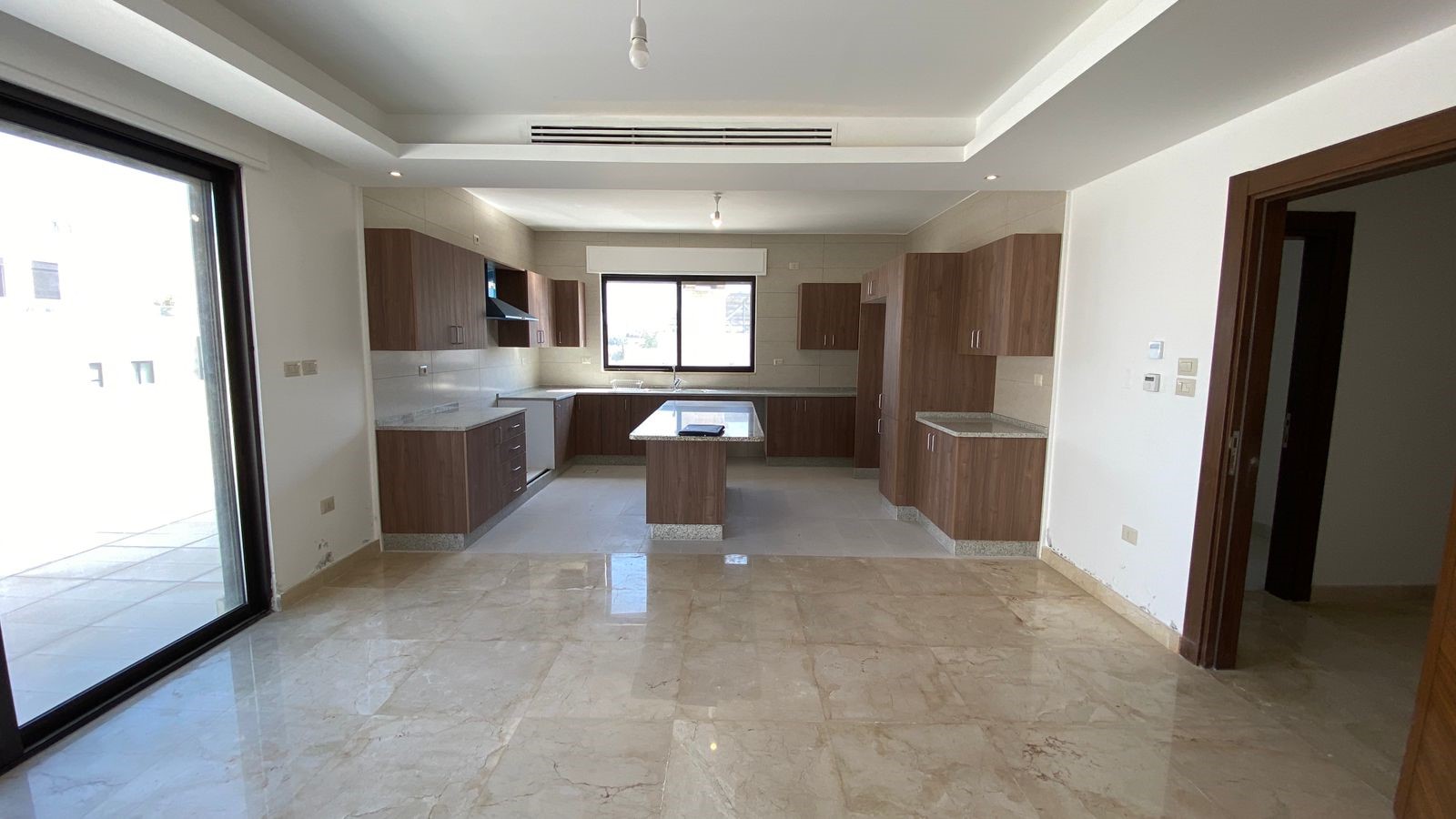 1fe4fb5d-8401-4b0b-b222-8b15f9621c1d Open-plan kitchen with wooden cabinets, central island, and marble floor; large sliding glass door to a patio on the left.