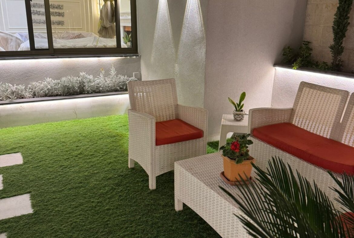 Outdoor patio with white wicker chairs and sofa with orange cushions on artificial grass, a white coffee table, and potted plants nearby.