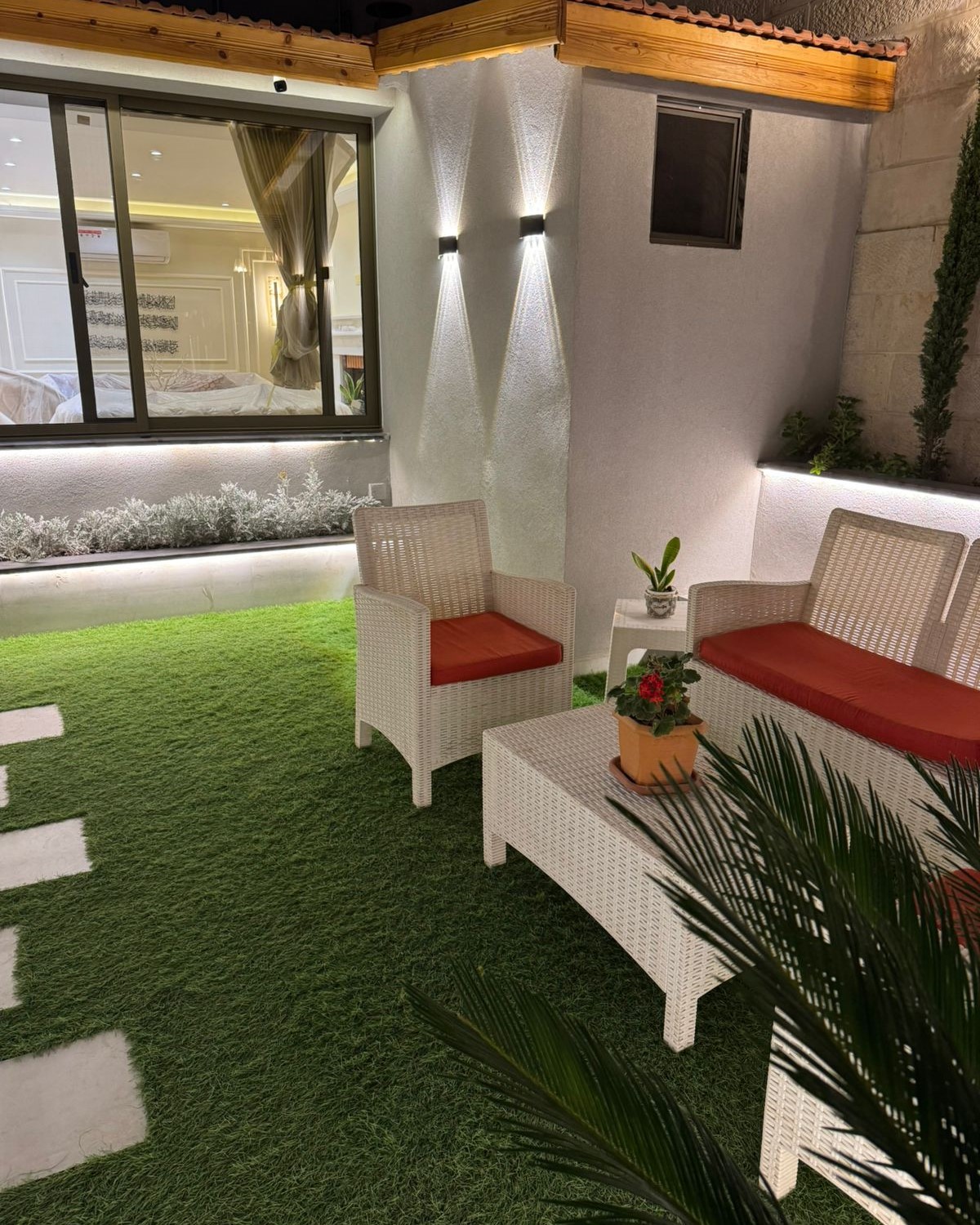 21ef74b0-8279-4405-b8d7-76803ad94087 Outdoor patio with white wicker chairs and sofa with orange cushions on artificial grass, a white coffee table, and potted plants nearby.
