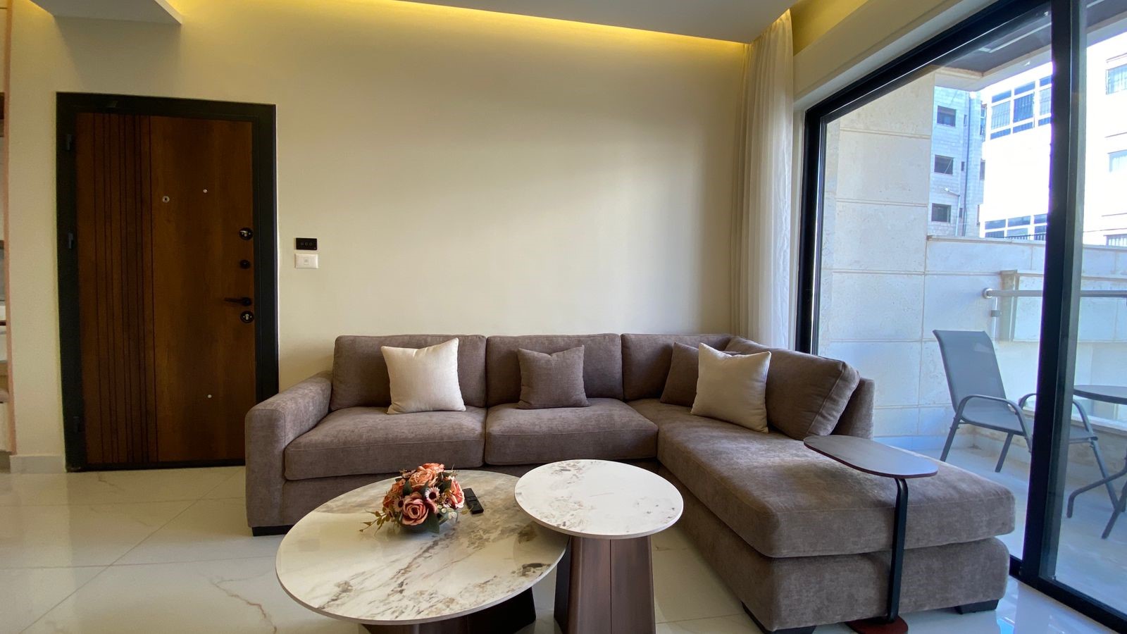 230bf90f-0d46-4a08-80d2-7e20115ecb08 L-shaped beige sofa with cream and brown cushions in a bright living room, marble coffee tables, and a balcony door on the right.