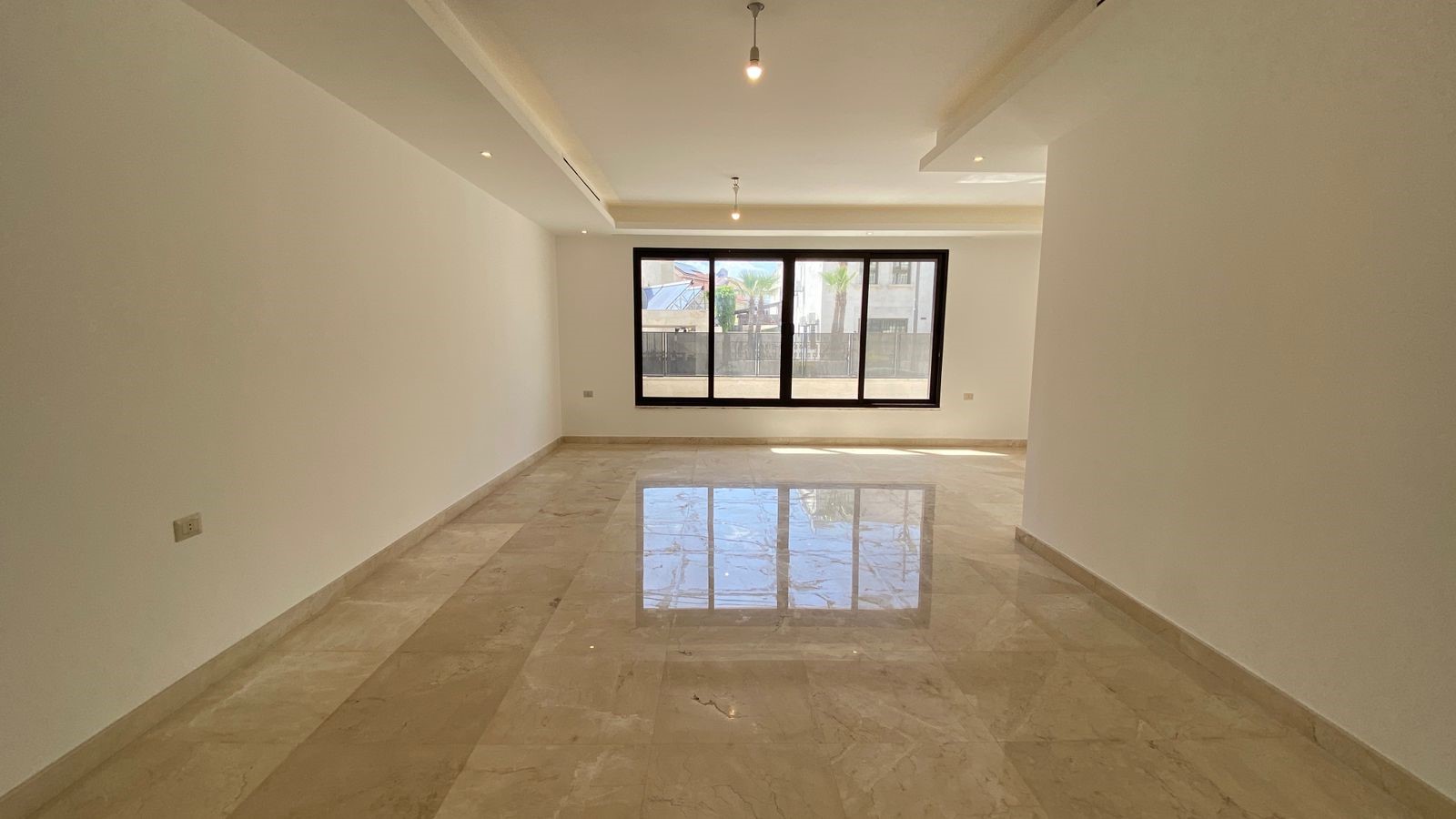 231f7ded-268e-44aa-abba-145c458c48bb Empty living room with a large dark-framed window, glossy beige marble floor, and white walls. Ceiling lights hang from above.