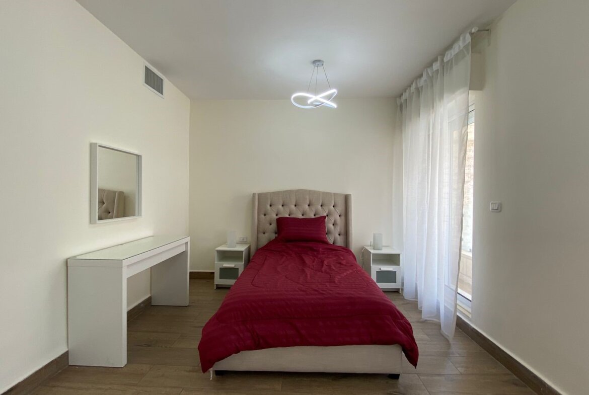 Bedroom with a single bed featuring a tufted beige headboard, red duvet, and white nightstands on either side. A white dresser with a mirror is against the wall, and a modern loop ceiling light hangs above.