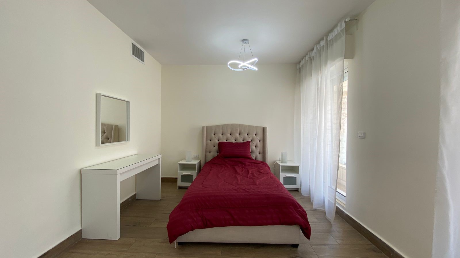 23ec51c7-be1e-4989-819b-9a9be268154c Bedroom with a single bed featuring a tufted beige headboard, red duvet, and white nightstands on either side. A white dresser with a mirror is against the wall, and a modern loop ceiling light hangs above.