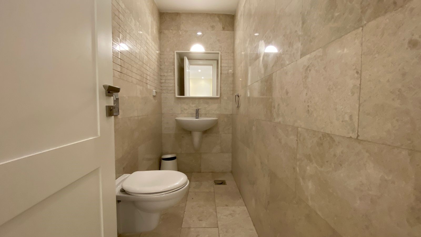 252b61ba-c467-4494-b33a-5b8d44ff1938 Narrow beige marble bathroom with a wall-mounted sink and mirror at the far end; toilet on the left and a small trash can nearby.