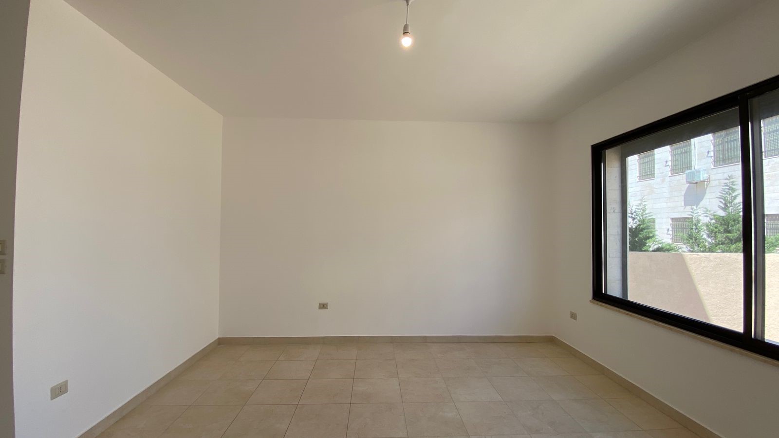 25922cdb-8065-401f-87f1-c5e47fcc5834 Empty room with beige floor tiles, white walls, a single bare light bulb, and a large right-side window letting in daylight.