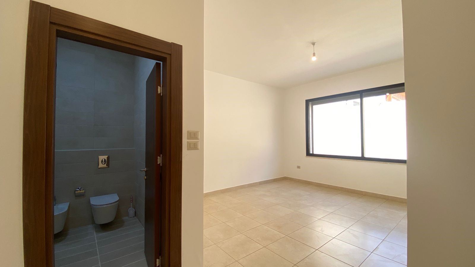 25c3ffc5-eae9-4efe-ba3e-9cc2d5eb6e21 Empty living room with beige walls, a large window, and beige tiled floor; doorway opens to a blue-tiled bathroom on the left.