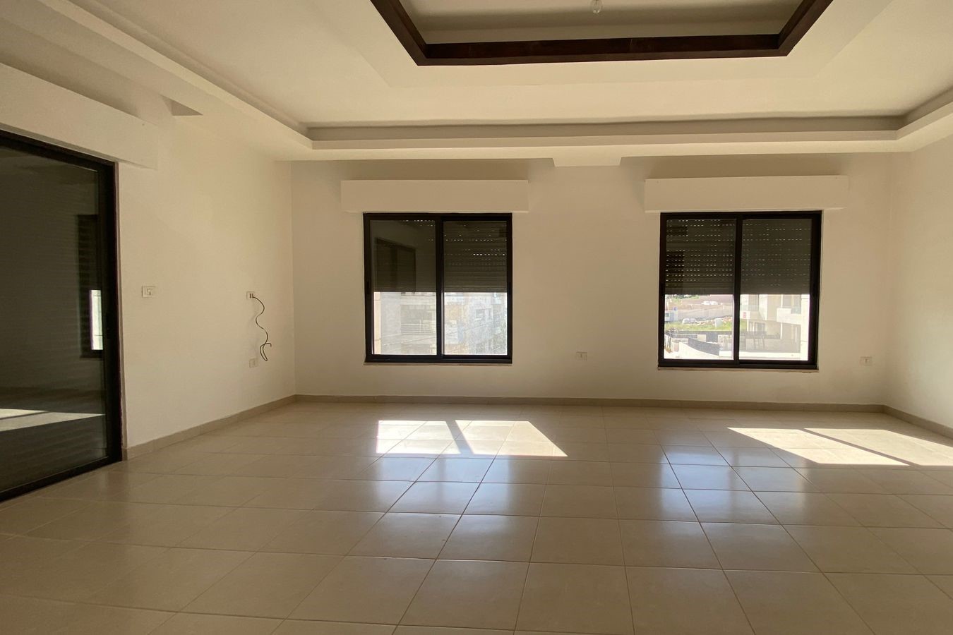 260492e8-8ac0-41c5-9c6d-c0f1047db633 Empty living room with beige tiled floor, cream walls, and two large windows letting in sunlight.