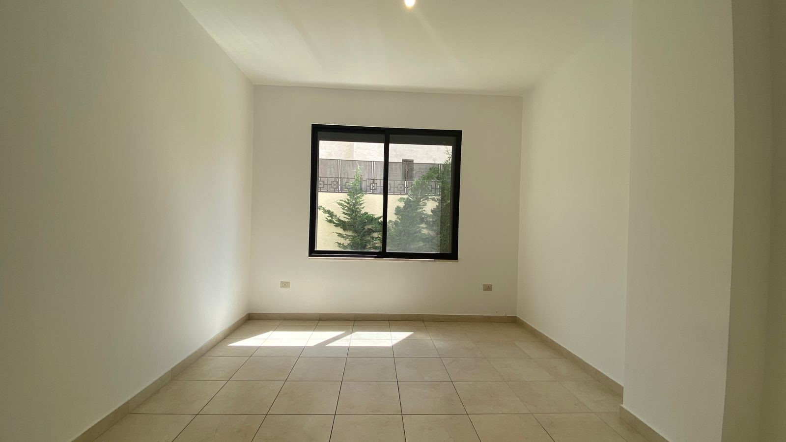26536738-d1d9-492d-85c9-325ad9229e44 Empty white room with a black-framed window letting in sunlight, beige tiled floor, blank walls.