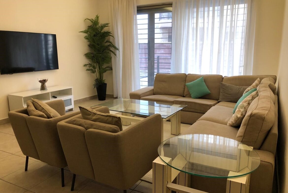 Bright modern living room with a beige sectional sofa, two armchairs, glass coffee table, and a wall-mounted TV; sheer white curtains over a balcony door in the background.